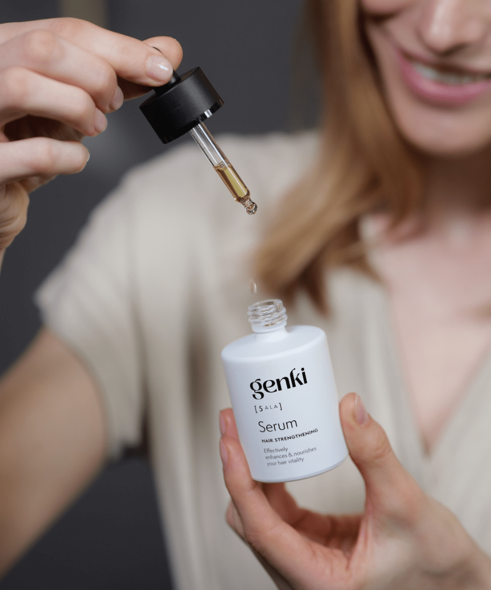 Nahaufnahme einer Frau, die mit einer Pipette eine goldene Flüssigkeit aus einer weißen Flasche mit der Aufschrift 'genki [5-ALA] Serum – Hair Strengthening' entnimmt. Das Serum tropft zurück in die Flasche. Die Frau lächelt dezent im Hintergrund