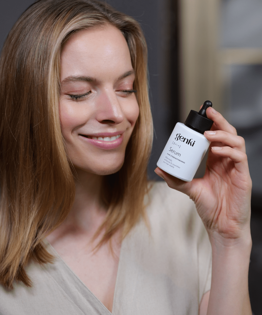 Eine lächelnde Frau mit schulterlangem, gesund glänzendem Haar hält eine weiße Flasche des 'genki [5-ALA] Hair Strengthening Serum' in der Hand. Sie hat die Augen geschlossen und wirkt entspannt, während sie das Produkt präsentiert.