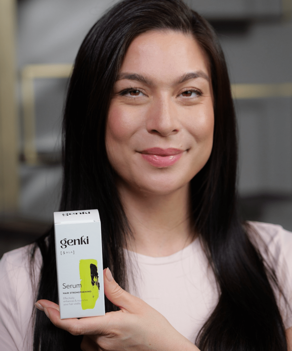 Eine Frau mit langem, glänzendem dunklem Haar lächelt sanft und hält die Verpackung des 'genki [5-ALA] Hair Strengthening Serum' in die Kamera. Die stilvolle weiße Schachtel mit schwarzer und gelber Gestaltung hebt sich klar hervor.